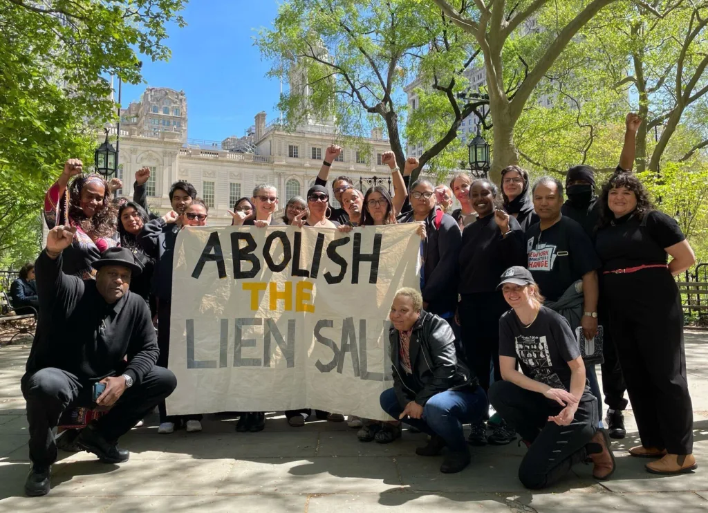A group of people rally outside of New York City Hall, holding a large sign reading "Abolish the Lien Sale."